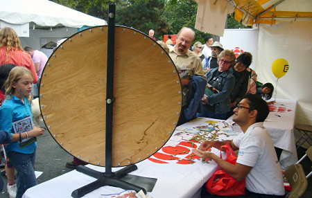 CBC table at outdoor fair has large vertical spinning wheel, its blank plywood back visible