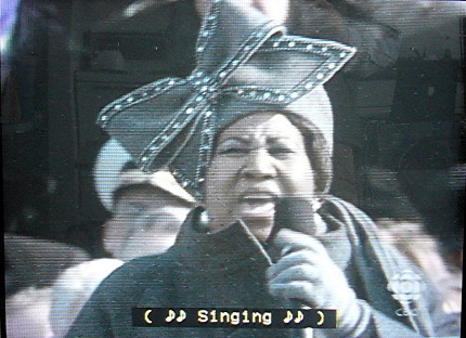 Aretha Franklin wears a grey outfit with matching hat and huge bow. Caption reads ( ♪♪ Singing ♪♪ )